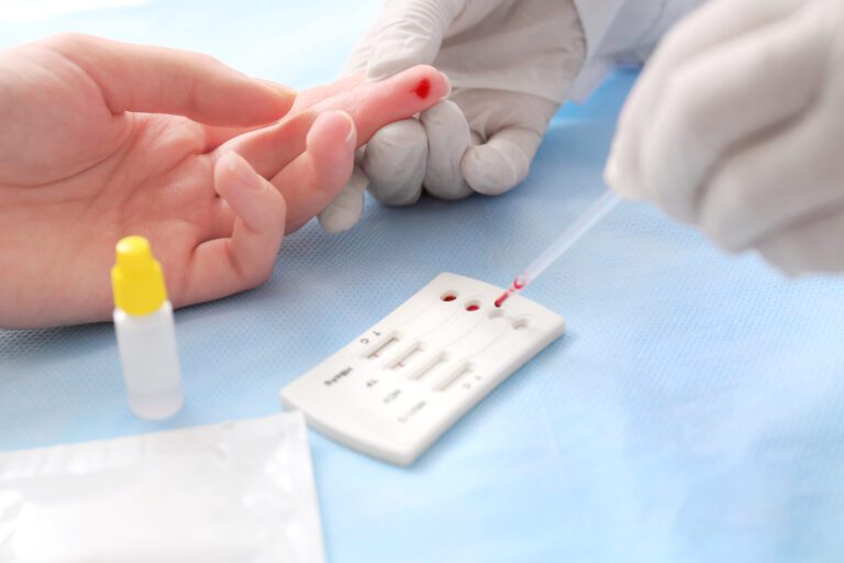 Pessoa com luvas nas mãos realizando o teste de Infecções sexualmente transmissíveis em outra pessoa. Aparecem apenas as mãos de ambas, e aparece o momento em que o sangue é coletado e é colocado na placa do teste.