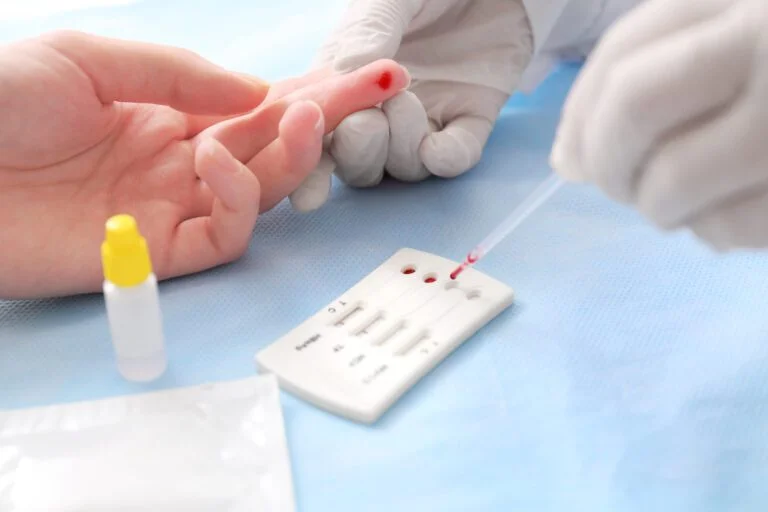 Pessoa com luvas nas mãos realizando o teste de Infecções sexualmente transmissíveis em outra pessoa. Aparecem apenas as mãos de ambas, e aparece o momento em que o sangue é coletado e é colocado na placa do teste.